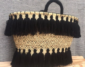 Puede incluir: Un bolso tote de paja tejido con ribete de borlas negras. El bolso tiene dos borlas negras a cada lado y un asa negra.