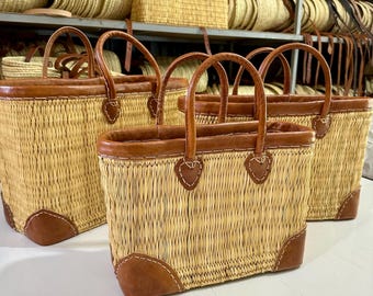 May include: Several rectangular woven straw tote bags with brown leather trim and handles. The bags have a light tan color and are displayed in a shop with other woven items on shelves in the background.