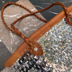 K&ouml;nnte beinhalten: Eine silberne Pailleten-Tragetasche mit einem geflochtenen braunen Ledergriff. Die Tasche hat einen braunen Lederbesatz und ist mit zahlreichen gl&auml;nzenden, runden Pailletten verziert. Der Griff ist mit braunem Leder und Metallnieten befestigt.