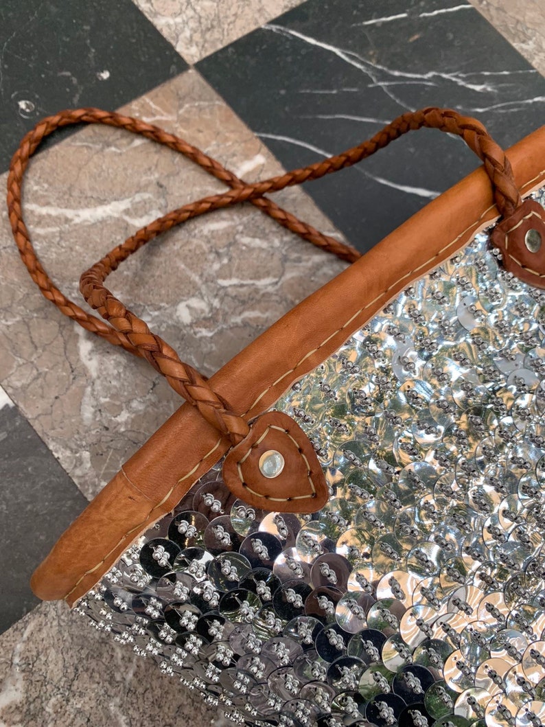 K&ouml;nnte beinhalten: Eine silberne Pailleten-Tragetasche mit einem geflochtenen braunen Ledergriff. Die Tasche hat einen braunen Lederbesatz und ist mit zahlreichen gl&auml;nzenden, runden Pailletten verziert. Der Griff ist mit braunem Leder und Metallnieten befestigt.