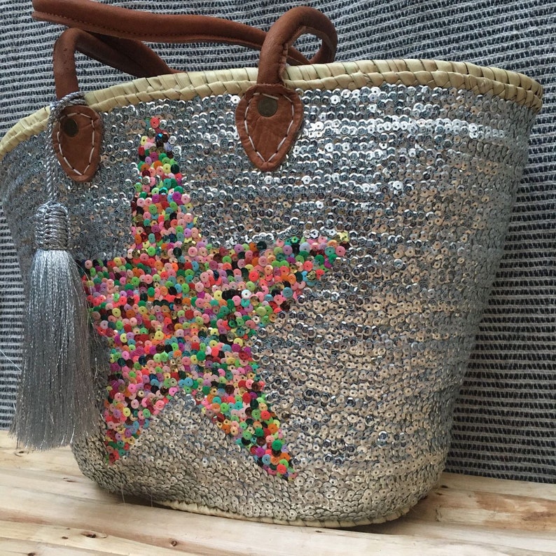 May include: A silver sequined tote bag with a star design made of colorful beads. The bag has brown leather handles and a silver tassel. The tote is made of woven straw.