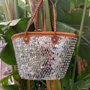 K&ouml;nnte beinhalten: Eine silberne Pailleten-Tragetasche mit braunen geflochtenen Griffen und braunem Wildlederbesatz. Die Tasche ist mit zahlreichen gl&auml;nzenden Pailletten verziert, die eine strukturierte Oberfl&auml;che erzeugen. Die Griffe sind aus geflochtenem Leder.