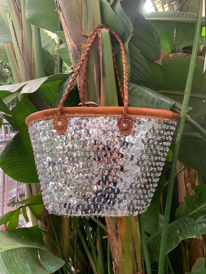 K&ouml;nnte beinhalten: Eine silberne Pailleten-Tragetasche mit braunen geflochtenen Griffen und braunem Wildlederbesatz. Die Tasche ist mit zahlreichen gl&auml;nzenden Pailletten verziert, die eine strukturierte Oberfl&auml;che erzeugen. Die Griffe sind aus geflochtenem Leder.