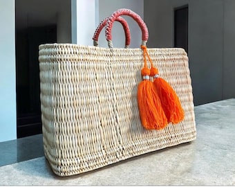 May include: A rectangular woven straw tote bag with coral-colored handles and orange tassels. The bag has a natural beige color and is designed for carrying items. The handles are wrapped with a coral-colored material.