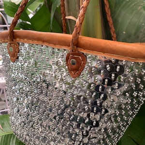 K&ouml;nnte beinhalten: Eine silberne Pailleten-Tragetasche mit braunem Lederbesatz und geflochtenen Henkeln. Die Tasche ist mit gl&auml;nzenden, runden Pailletten bedeckt, die eine strukturierte, reflektierende Oberfl&auml;che bilden. Die Henkel und der Besatz sind warmbraun.