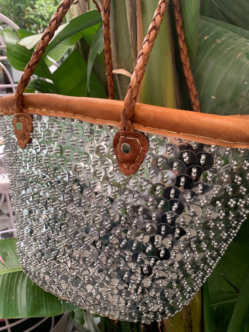 K&ouml;nnte beinhalten: Eine silberne Pailleten-Tragetasche mit braunem Lederbesatz und geflochtenen Henkeln. Die Tasche ist mit gl&auml;nzenden, runden Pailletten bedeckt, die eine strukturierte, reflektierende Oberfl&auml;che bilden. Die Henkel und der Besatz sind warmbraun.