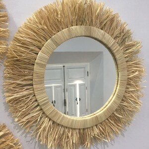 May include: A round mirror with a natural woven frame. The frame is made of straw and has a fringe around the edge. The mirror reflects a white door with a window.