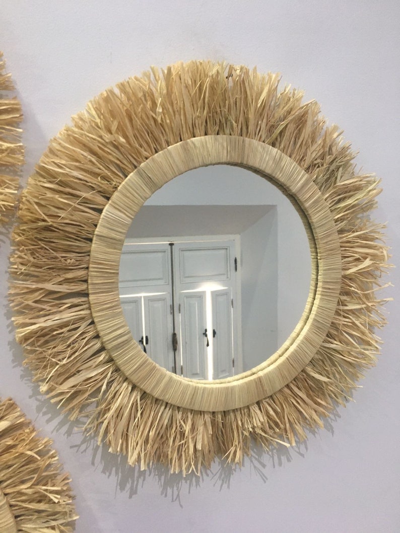 May include: A round mirror with a natural woven frame. The frame is made of straw and has a fringe around the edge. The mirror reflects a white door with a window.