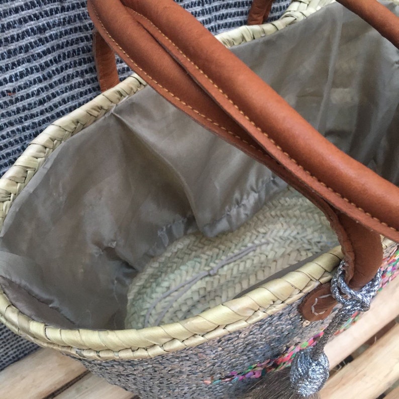 May include: Woven straw tote bag with brown leather handles and a gray interior lining. The bag features a decorative tassel and colorful accents. A stylish and practical accessory for carrying items.