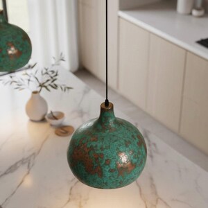 Puede incluir: L&aacute;mpara colgante esf&eacute;rica verde turquesa con un patr&oacute;n moteado de color cobre. La l&aacute;mpara cuelga de un cable negro con un accesorio de lat&oacute;n. Se suspende sobre una encimera de m&aacute;rmol en una cocina.