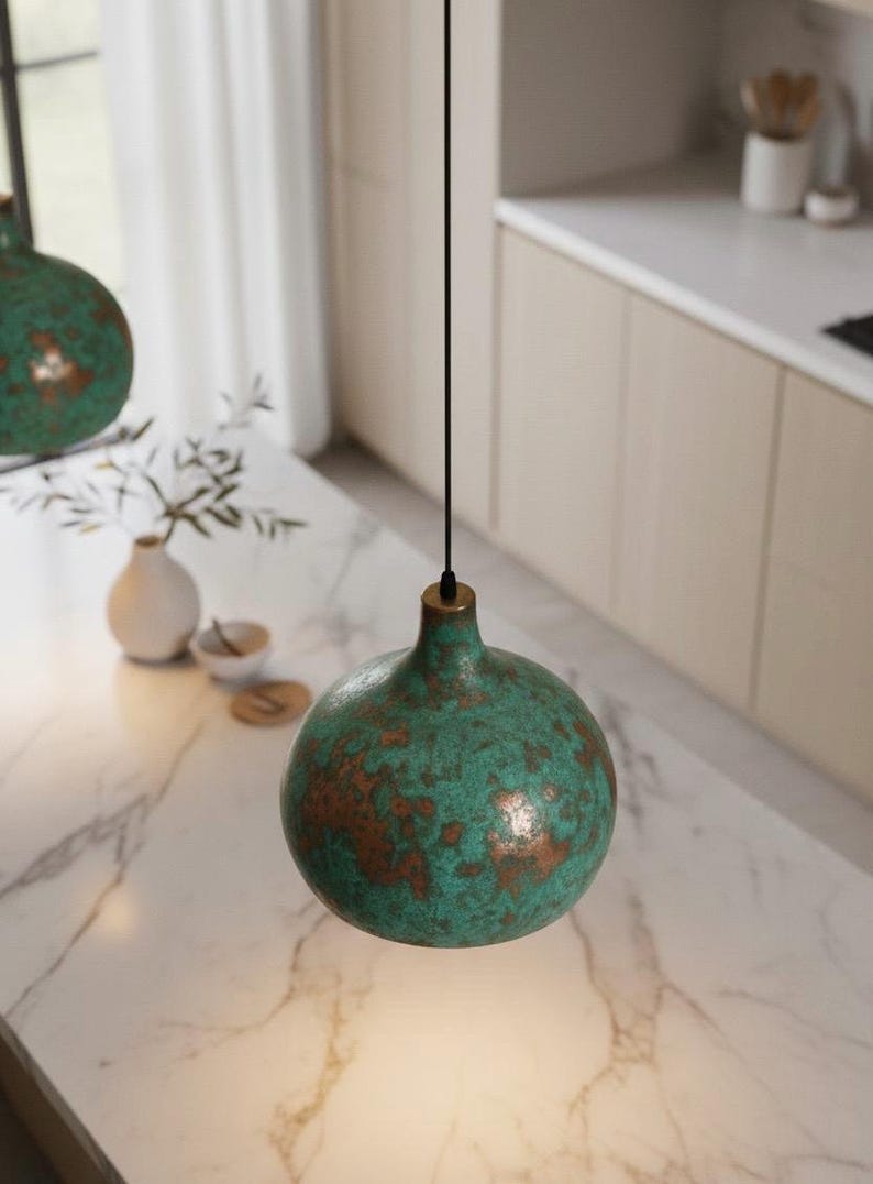 Puede incluir: L&aacute;mpara colgante esf&eacute;rica verde turquesa con un patr&oacute;n moteado de color cobre. La l&aacute;mpara cuelga de un cable negro con un accesorio de lat&oacute;n. Se suspende sobre una encimera de m&aacute;rmol en una cocina.