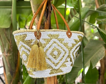 Peut inclure: Un sac cabas en perles blanches et dorées avec un motif géométrique et un pompon doré. Le sac a des anses en cuir marron.