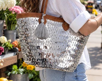 May include: A silver sequin tote bag featuring a brown leather trim, brown leather shoulder straps, and a decorative grey tassel attached to the top. The bag is covered in reflective circular sequins that create a shimmering metallic appearance.