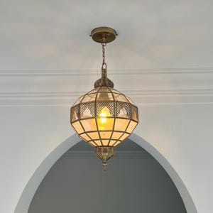 May include: Ornate, antique-style pendant light fixture. The brass-colored metal frame features a geometric design with glass panels. The light is suspended from a chain and emits a warm, inviting glow. The fixture is a decorative element.