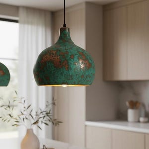 Puede incluir: Una l&aacute;mpara colgante con acabado verde y cobre. La l&aacute;mpara tiene forma redondeada y cuelga de un cable negro. El accesorio est&aacute; en un entorno de cocina con una encimera blanca y un jarr&oacute;n con vegetaci&oacute;n.