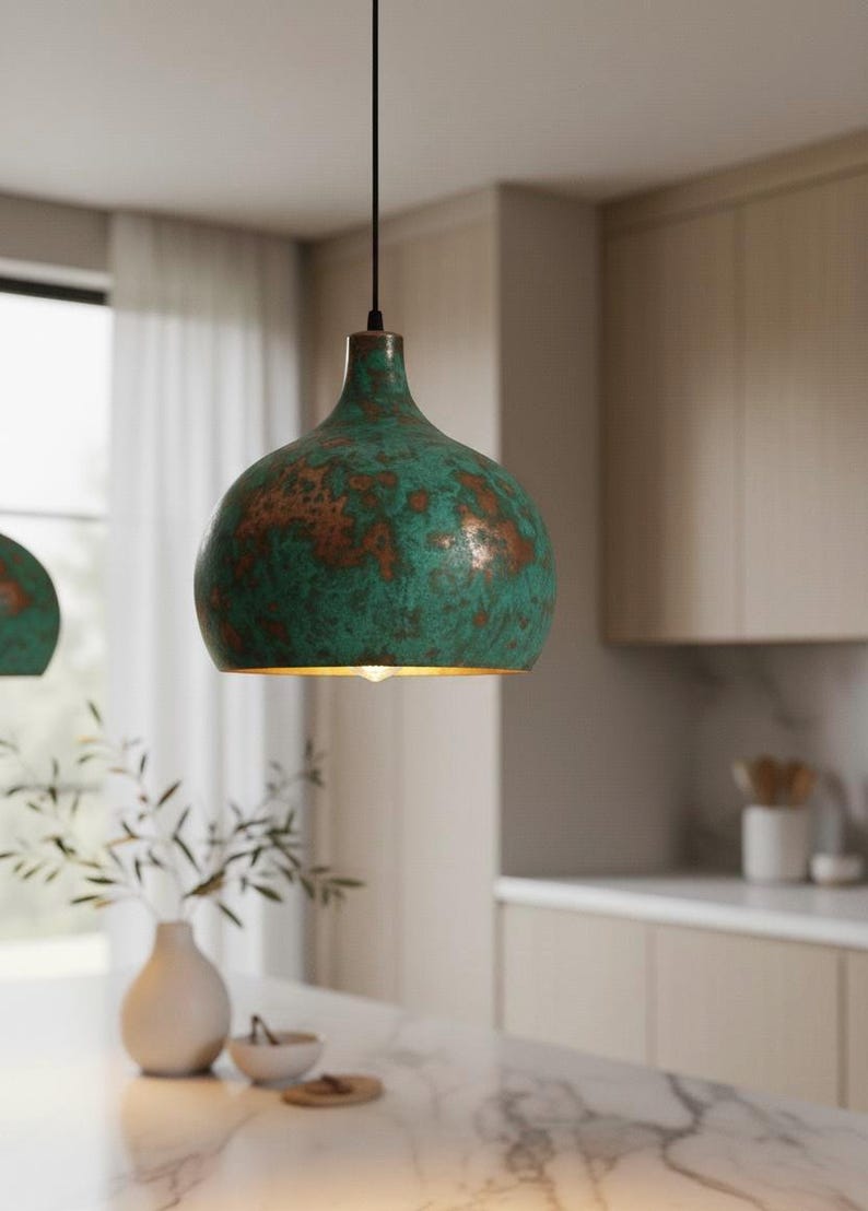 Puede incluir: Una l&aacute;mpara colgante con acabado verde y cobre. La l&aacute;mpara tiene forma redondeada y cuelga de un cable negro. El accesorio est&aacute; en un entorno de cocina con una encimera blanca y un jarr&oacute;n con vegetaci&oacute;n.