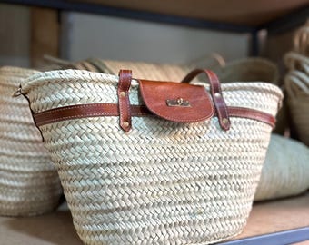 May include: Woven straw tote bag with brown leather accents. The bag features a brown leather flap with a metal clasp closure and a brown leather handle. The bag is sitting on a shelf with other similar bags in the background.