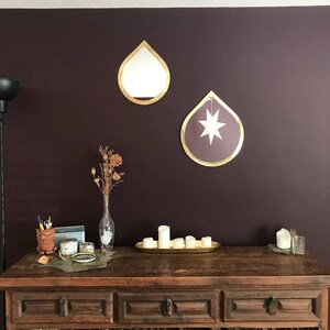 May include: Two gold teardrop-shaped mirrors hang on a dark purple wall above a wooden table with drawers. The mirror on the left is plain, while the mirror on the right has a white star inside.