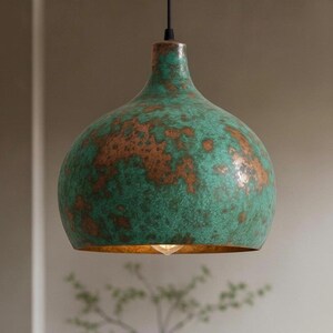 Puede incluir: L&aacute;mpara colgante con acabado verde y cobre. La l&aacute;mpara tiene una pantalla redondeada en forma de campana con una superficie texturizada. El interior de la pantalla es de color dorado y se ve una sola bombilla.