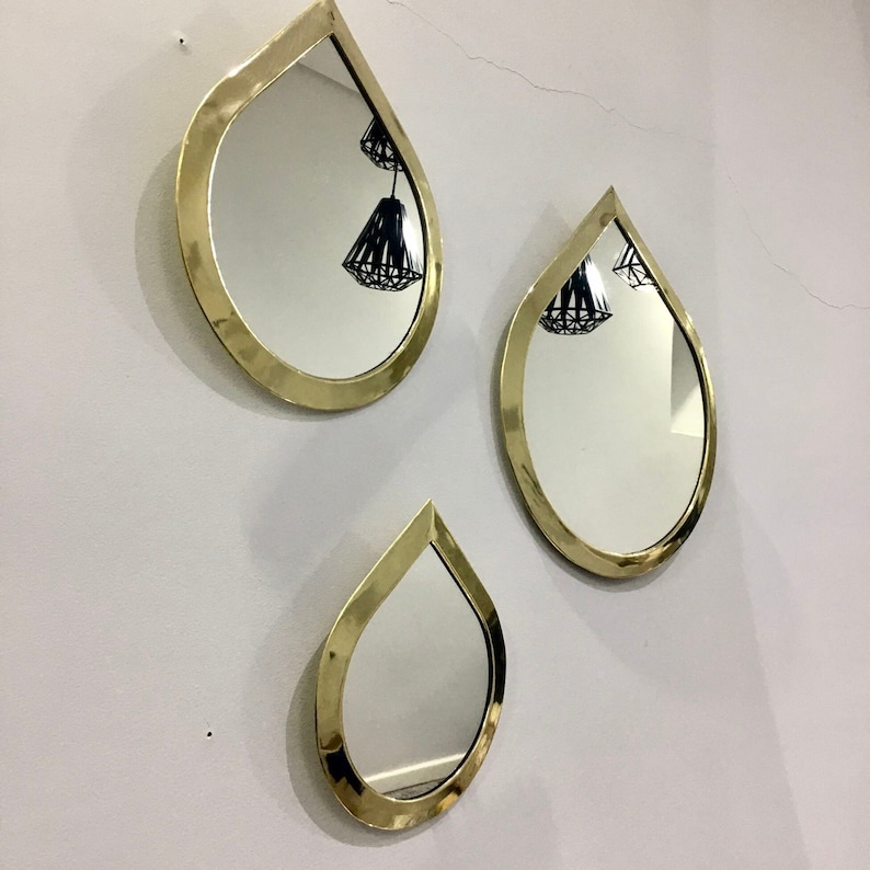 May include: Three gold teardrop-shaped mirrors with a reflective surface. The mirrors are hanging on a white wall.