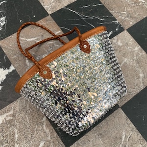 K&ouml;nnte beinhalten: Eine silberne Pailleten-Tragetasche mit braunen Lederbes&auml;tzen und geflochtenen Griffen. Die Tasche ist rechteckig mit abgerundeter Oberseite und besteht aus gl&auml;nzenden, reflektierenden Pailletten. Die Griffe sind hellbraun.