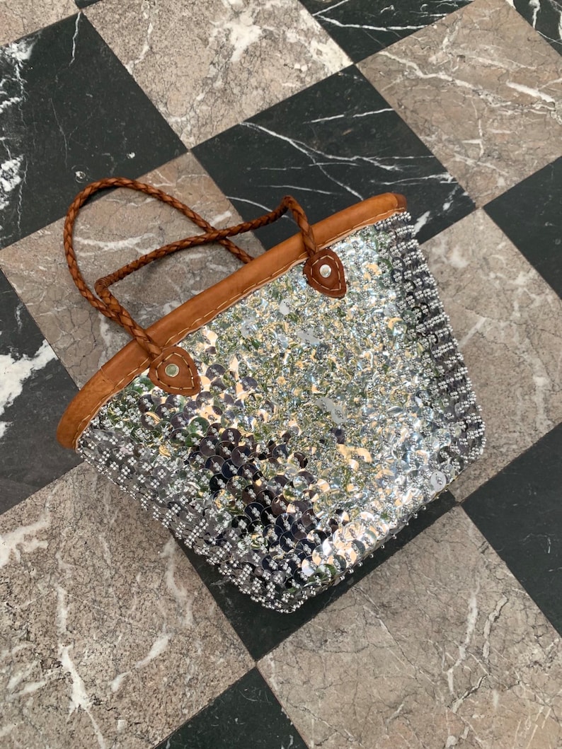 K&ouml;nnte beinhalten: Eine silberne Pailleten-Tragetasche mit braunen Lederbes&auml;tzen und geflochtenen Griffen. Die Tasche ist rechteckig mit abgerundeter Oberseite und besteht aus gl&auml;nzenden, reflektierenden Pailletten. Die Griffe sind hellbraun.