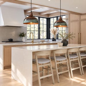May include: Two dome-shaped pendant lights with a weathered copper and teal patina finish hang above a kitchen island. The island features a white countertop and five light-colored wooden stools with grey upholstered seats in a kitchen.
