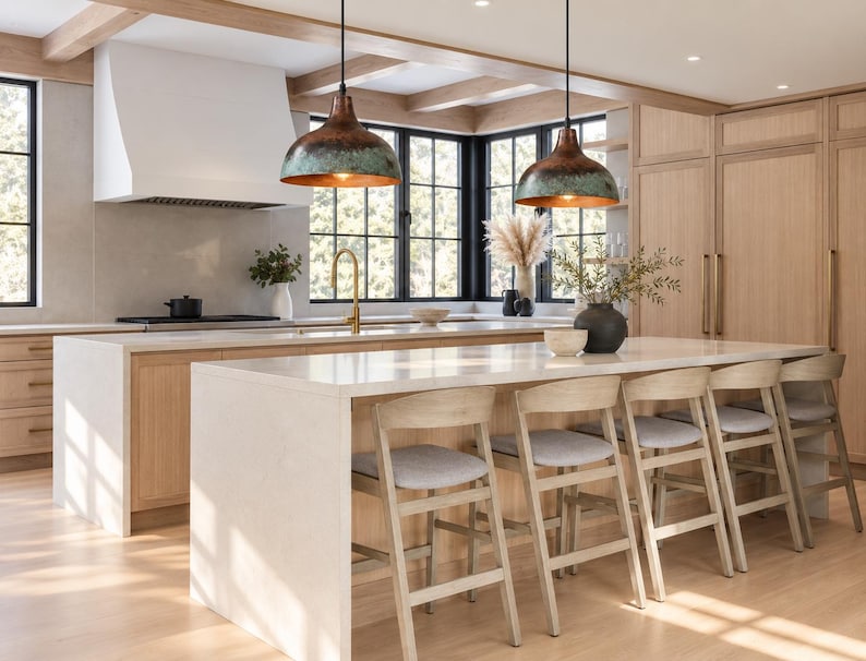 May include: Two dome-shaped pendant lights with a weathered copper and teal patina finish hang above a kitchen island. The island features a white countertop and five light-colored wooden stools with grey upholstered seats in a kitchen.