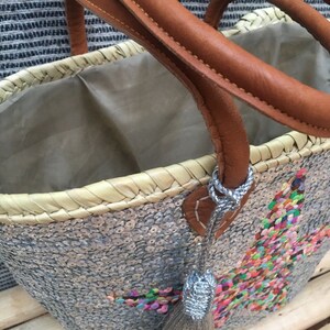 May include: A silver sequined tote bag with brown leather handles and a colorful embroidered design. The bag has a woven straw base and a silver tassel. The interior is lined with a light brown fabric.