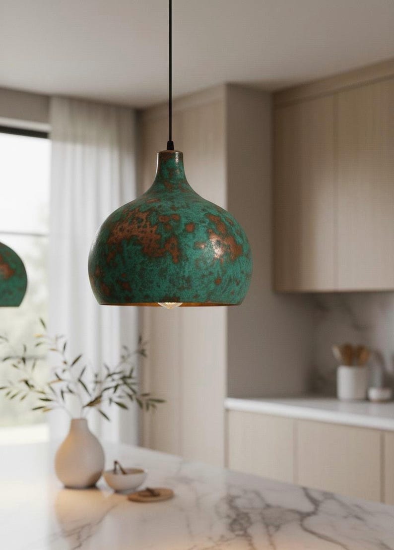 Puede incluir: Una l&aacute;mpara colgante verde y cobriza de forma redondeada cuelga de un cable negro. La luz tiene un acabado envejecido y est&aacute; suspendida sobre una encimera de cocina. El accesorio es un elemento decorativo.