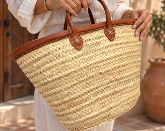 May include: A woven straw basket bag featuring a light beige natural fiber body with a chevron pattern. The bag includes two brown leather handles attached with reinforced leather patches and silver-toned metal rivets at the top rim.