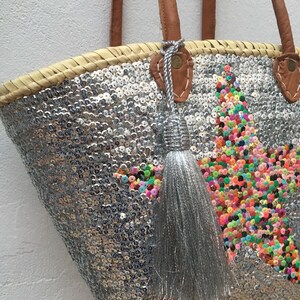 May include: A silver sequined tote bag with a star design made of colorful sequins. The bag has brown leather handles and a silver tassel. The bag is made of woven straw.