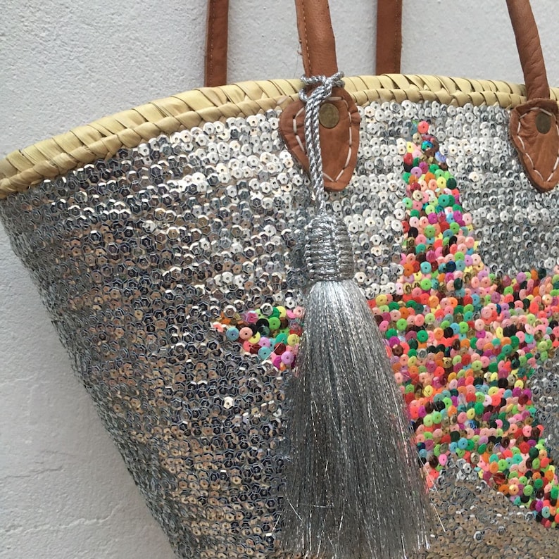 May include: A silver sequined tote bag with a star design made of colorful sequins. The bag has brown leather handles and a silver tassel. The bag is made of woven straw.