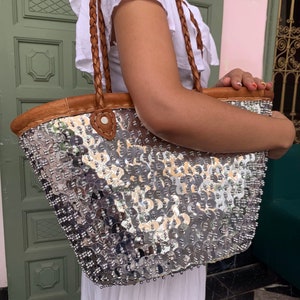 May include: A silver sequined tote bag with brown leather trim and braided brown leather handles. The bag is covered in shiny, round sequins and has a structured shape. The handles are thick and appear sturdy. The bag is a fashionable accessory.