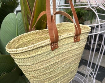May include: A large, light brown woven basket with two long brown leather straps. The basket is hanging from a tree branch.