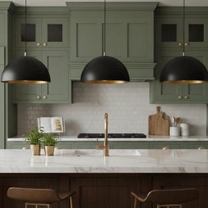 May include: Three black pendant lights with gold interiors hang above a kitchen island. The kitchen features sage green cabinets, a white marble countertop, and a dark wood base. A gold faucet and white subway tile backsplash complete the design.