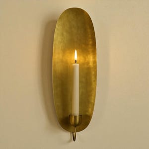 May include: A brass wall-mounted candle holder with an oval design. A lit white candle sits in the holder, casting a warm glow. The holder is attached to the wall. The candle holder is approximately 30 cm in height.