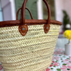 May include: A woven straw tote bag with brown leather handles and trim. The bag has a rounded shape and is sitting on a tiled surface.