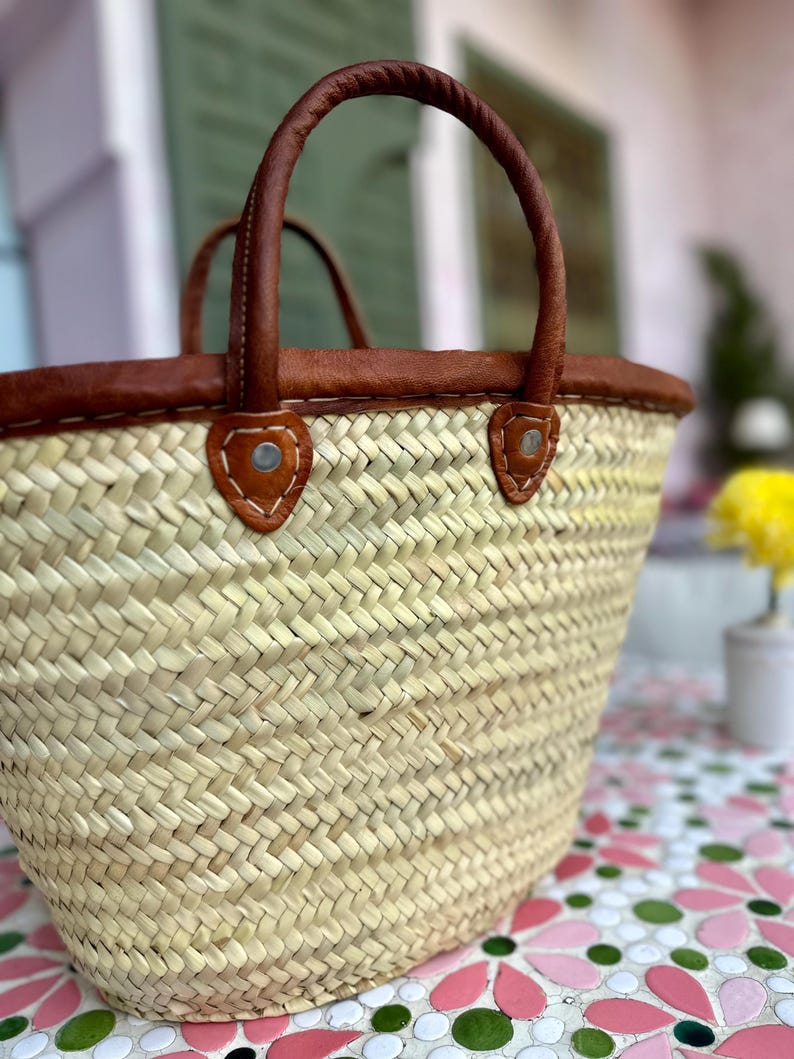 May include: A woven straw tote bag with brown leather handles and trim. The bag has a rounded shape and is sitting on a tiled surface.