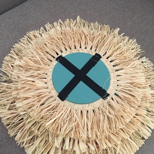 May include: A round wall decor with a woven straw fringe. The center of the decor is a teal blue circle with a black X across it.