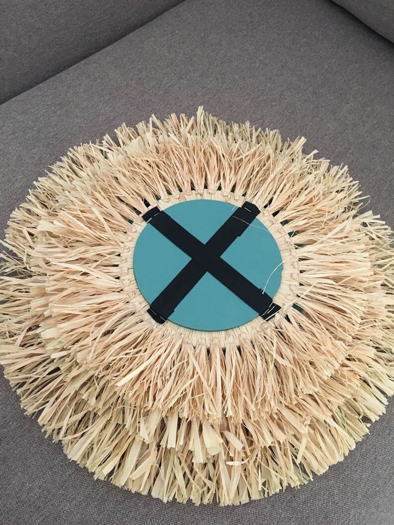 May include: A round wall decor with a woven straw fringe. The center of the decor is a teal blue circle with a black X across it.