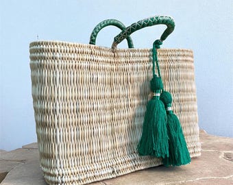 May include: A rectangular woven straw tote bag with green leather handles and a green tassel. The bag is made of natural-colored straw and has a structured shape. The handles are curved and stitched with white thread.