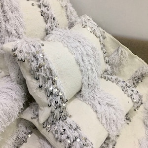 May include: White throw pillows with fringe and silver sequins. The pillows are stacked on top of each other.