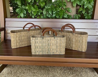 May include: Three woven rectangular baskets with brown leather handles. The baskets are made of light brown and tan woven material. The handles are a darker brown color. The baskets are sitting on a brown wooden bench.
