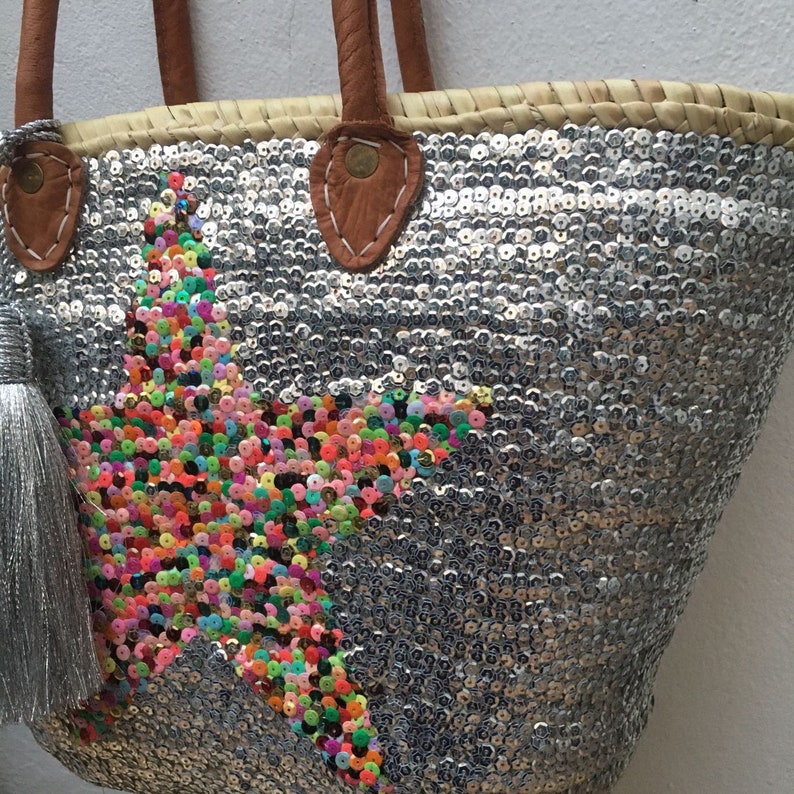 May include: A silver sequined tote bag with a star design made of colorful sequins. The bag has brown leather handles and a silver tassel. The bag is made of woven straw.