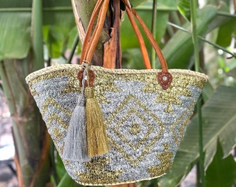 Peut inclure: Un sac cabas en sequins argentés et dorés avec des anses en cuir marron et un pompon. Le sac est suspendu à une branche d'arbre.