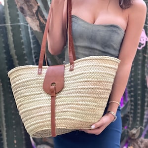 May include: A large, woven straw tote bag with brown leather handles and a brown leather strap closure. The bag is light beige in colour and has a woven pattern.