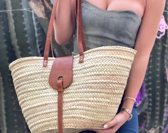 Pode incluir: Uma grande bolsa tote de palha tecida com alças de couro marrom e um fecho de tira de couro marrom. A bolsa é de cor bege claro e tem um padrão tecido.