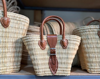 May include: Woven straw tote bags with brown leather handles and accents. The bags have a rectangular shape with a buckle closure and heart-shaped leather details. Several bags are visible, arranged on a shelf.