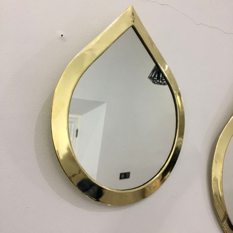 May include: A pair of gold teardrop-shaped mirrors with a smooth, reflective surface. The mirrors are mounted on a white wall.